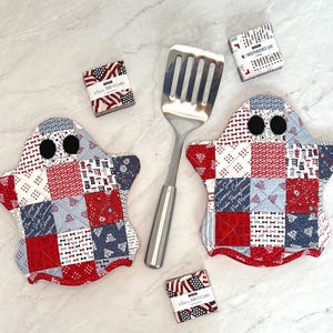 Puede incluir: Dos agarraderas acolchadas con forma de fantasma con un patrón de patchwork en rojo, blanco y azul y ojos circulares negros, una espátula ranurada de acero inoxidable y tres pequeños paquetes de tela cuadrados etiquetados Independence Day y America 250 Years.