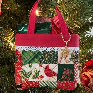 May include: A red quilted fabric tote bag with a white lace trim, a gold chain, and a gold snowflake charm. The bag is filled with wrapped chocolates and is hanging on a Christmas tree.