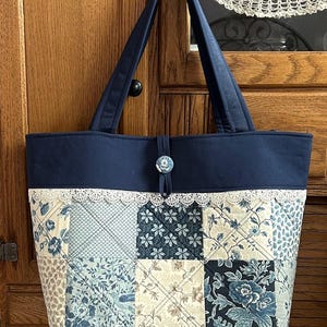 May include: A navy blue quilted tote bag with a patchwork design of blue and white floral fabric. The bag has a white lace trim and a blue button closure.