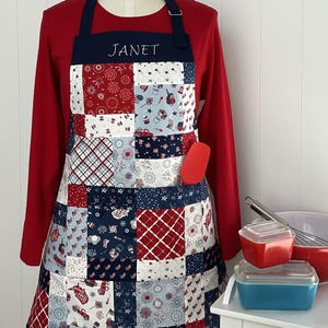 Op de afbeelding: Een patriottische patchwork schort met rode, witte en blauwe patronen, met vuurwerk, vlaggen en sterren. De schort heeft een marineblauwe nekband en zak, met de naam "JANET" geborduurd op de slab. Een rode spatel zit in de zak.