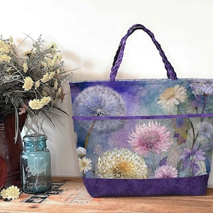 May include: A floral tote bag with a purple braided handle. The bag features a watercolour design of dandelions and other flowers in shades of purple, pink, and yellow. The bottom and trim are a solid purple fabric.