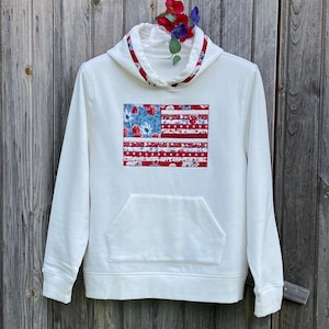 May include: White hooded sweatshirt with a red, white, and blue American flag design made of fabric scraps. The flag is centered on the front of the sweatshirt.