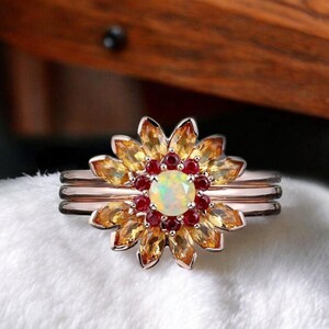 May include: A gold ring featuring a white opal centre stone surrounded by smaller red and yellow gemstones, creating a flower design.