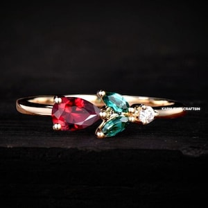 Op de afbeelding: Een gouden ring met een rode peervormige edelsteen, twee groene peervormige edelstenen en een kleine ronde witte edelsteen. De ring ligt op een zwart oppervlak.