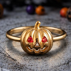 Op de afbeelding: Een goudkleurige ring met een centrale bedel in de vorm van een pompoen. De pompoen heeft een uitgesneden gezicht met twee kleine, fonkelende rode edelstenen die in de driehoekige oogopeningen zijn gezet, wat zorgt voor een helder contrast.
