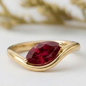 Könnte beinhalten: Ein goldener Ring mit einem marquiseförmigen, tiefroten Edelstein. Das Band des Rings hat ein fließendes, wellenartiges Design. Der Edelstein ist der Mittelpunkt, gefasst in einer polierten Goldfassung. Der Hintergrund ist unscharf.
