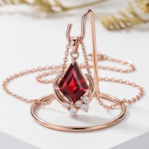 Op de afbeelding: Een roségouden ketting met een vliegervormige rode edelsteen hanger. De hanger is versierd met een halve maan vorm en kleine heldere steentjes aan de onderkant. De fijne roségouden ketting hangt aan een gebogen metalen displaystandaard.