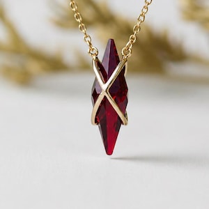 Puede incluir: Un collar de cadena dorada con un colgante de cristal rojo intenso, facetado y puntiagudo. El cristal se sujeta mediante un delicado engaste de metal dorado cruzado que asegura la piedra en el centro, creando una forma geométrica alargada.