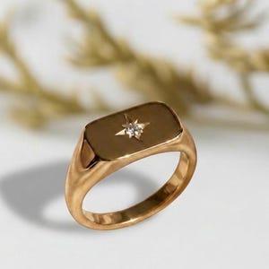 May include: A gold-colored signet ring with a rectangular face. The ring features a starburst design with a small, clear gemstone at its center. The ring is set against a white background with a blurred, golden-colored plant in the background.