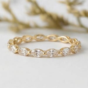 May include: A gold eternity band featuring a continuous row of marquise-cut clear stones set in a polished yellow gold metal frame. The ring displays a repeating pattern of faceted gems held by small gold prongs, creating a delicate and uniform circular design.