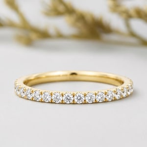 Peut inclure: Une bague en or avec une rangée continue de petites pierres précieuses rondes et claires. La bague est posée sur un fond doux et neutre avec un feuillage flou de couleur dorée.