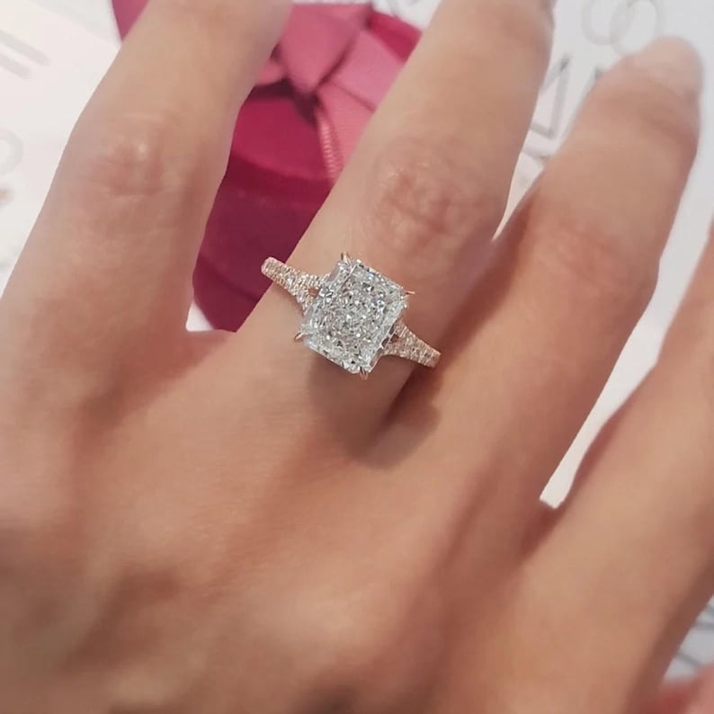 Puede incluir: Un anillo de compromiso de oro con un gran diamante de forma cuadrada rodeado de diamantes m&aacute;s peque&ntilde;os. El anillo est&aacute; en un dedo.