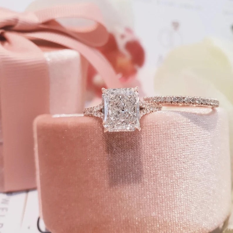 Puede incluir: Primer plano de un anillo de compromiso de diamante de forma cuadrada con una banda delicada. El anillo est&aacute; engastado en oro rosa y est&aacute; rodeado de diamantes m&aacute;s peque&ntilde;os. El anillo est&aacute; sentado en una caja de terciopelo rosa.