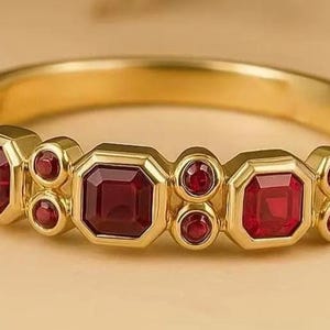 Op de afbeelding: Een gouden ring met afwisselend vierkante en ronde rode edelstenen. De ring heeft een gepolijste gouden band. De edelstenen zijn dieprood en geplaatst tegen een neutrale achtergrond.