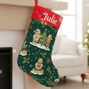 Puede incluir: Un calcetín navideño con puño rojo y cuerpo verde. El calcetín presenta un patrón festivo de ranas con suéteres y gorros de Papá Noel, decorando un árbol de Navidad. El nombre "Julie" está impreso en el puño.