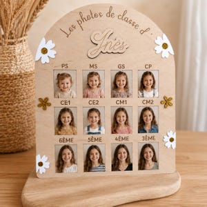 Op de afbeelding: Een boogvormig houten displaybord met de tekst Les photos de classe d' In&egrave;s, voorzien van twaalf rechthoekige vakjes voor schoolfoto's met labels van PS tot 3&Egrave;ME, versierd met witte en goudkleurige glitterbloemen van hout.