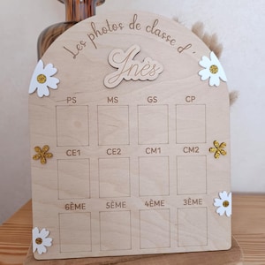 Op de afbeelding: Een boogvormig houten bord voor schoolfoto's met de tekst Les photos de classe d' In&egrave;s. Het bevat twaalf rechthoekige vakken gelabeld met Franse schoolniveaus van PS tot 3&Egrave;ME, versierd met witte en gouden glitterbloemen als decoratie.
