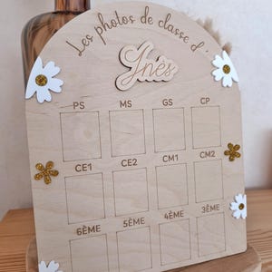 Op de afbeelding: Een boogvormig presentatiebord van licht hout met de tekst Les photos de classe d' In&egrave;s. Het bevat twaalf rechthoekige vakken gelabeld met schoolniveaus van PS tot 3&Egrave;ME, versierd met witte en gouden glitterbloemen als decoratie.