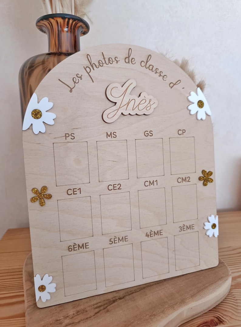 Op de afbeelding: Een boogvormig presentatiebord van licht hout met de tekst Les photos de classe d' In&egrave;s. Het bevat twaalf rechthoekige vakken gelabeld met schoolniveaus van PS tot 3&Egrave;ME, versierd met witte en gouden glitterbloemen als decoratie.