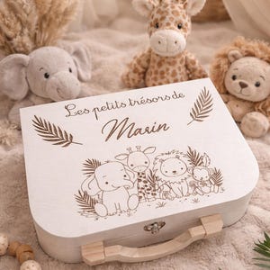 Peut inclure: Un coffret souvenir en bois blanc avec une poignée, orné d'une illustration gravée d'animaux de la jungle et du texte "Les petits trésors de Marin". Des peluches d'animaux, dont une girafe, un éléphant et un lion, sont disposées autour du coffret.