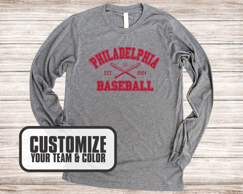 Puede incluir: Camiseta de manga larga gris con un dise&ntilde;o gr&aacute;fico rojo que dice "Philadelphia Baseball" con bates de b&eacute;isbol cruzados y una pelota de b&eacute;isbol. El texto "EST" y "2024" tambi&eacute;n est&aacute;n incluidos en el dise&ntilde;o. El texto "Personaliza tu equipo y color" est&aacute; en un cuadro blanco con borde negro.