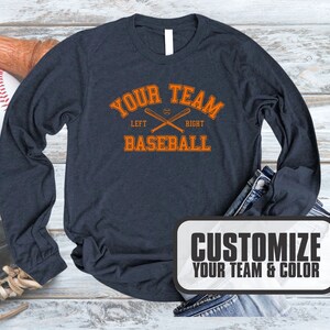 Puede incluir: Una camiseta de manga larga azul marino con un gr&aacute;fico de b&eacute;isbol naranja. El gr&aacute;fico dice "Your Team Baseball" con las palabras "Left" y "Right" debajo de los bates de b&eacute;isbol cruzados. El texto "Customize Your Team & Color" est&aacute; en un rect&aacute;ngulo blanco con borde negro.