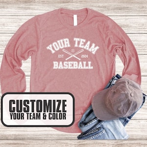 Puede incluir: Una camiseta rosa de manga larga con el texto "Your Team Baseball Est 2024" y un gr&aacute;fico de dos bates de b&eacute;isbol cruzados. La camiseta est&aacute; sobre un fondo de madera.