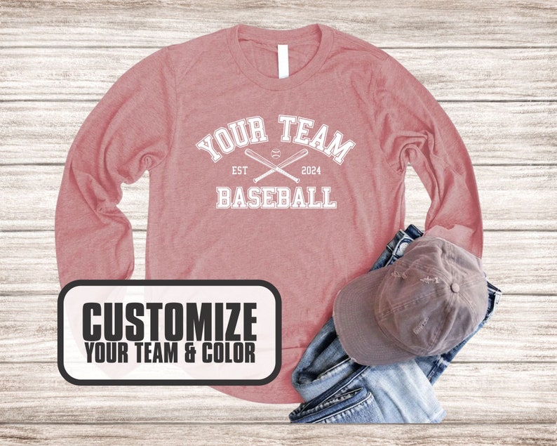 Puede incluir: Una camiseta rosa de manga larga con el texto "Your Team Baseball Est 2024" y un gr&aacute;fico de dos bates de b&eacute;isbol cruzados. La camiseta est&aacute; sobre un fondo de madera.