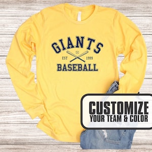 Puede incluir: Camiseta amarilla de manga larga con un gr&aacute;fico azul marino que dice "Giants Baseball Est 1999" con bates de b&eacute;isbol cruzados. El texto "Personaliza tu equipo y color" est&aacute; en un rect&aacute;ngulo blanco y negro.
