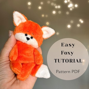 May include: A small plush fox toy with bright orange fur, a white muzzle, white inner ears, and a white tail tip. The fox has black embroidered eyes and a black nose. Next to the toy is a circular graphic with the text Easy Foxy TUTORIAL Pattern PDF.