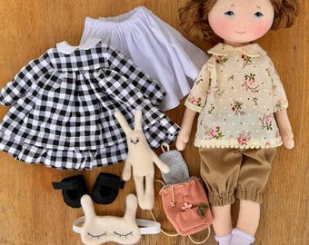 Op de afbeelding: Een handgemaakte pop met bruin haar en blauwe ogen, gekleed in een bloemen top en bruine broek. Accessoires zijn een zwart-wit geruite jurk, witte rok, zwarte schoenen, een konijn, een rugzak en een slaapmasker.