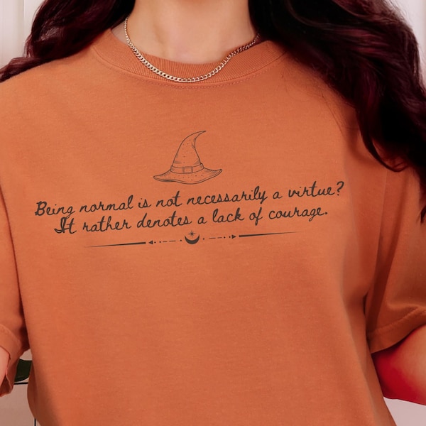 May include: A burnt orange crewneck t-shirt featuring a black graphic of a witch hat and the text: Being normal is not necessarily a virtue? It rather denotes a lack of courage. The design includes a small crescent moon and star symbol below.