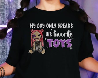 Op de afbeelding: Zwart T-shirt met een afbeelding van een gestikte pop met roze haar en de tekst "My boy only breaks his favorite toys".
