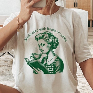 Op de afbeelding: Een beige T-shirt met een groene illustratie van een vrouw die een boek leest en koffie drinkt. De tekst "Medicated with books & coffee" staat op het T-shirt gedrukt.