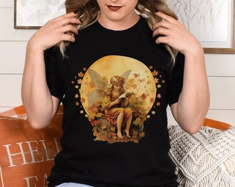 Op de afbeelding: Zwart T-shirt met een afbeelding van een fee die een boek leest in een cirkel van herfstbladeren.