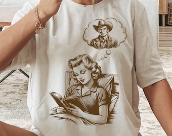May include: A woman wearing a white t-shirt with a brown illustration of a woman reading a book in bed and a thought bubble with a cowboy in it. The text in the thought bubble reads "oooo".