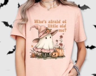 Op de afbeelding: Een roze T-shirt met een schattig spookje dat een heksenhoed draagt en een pompoen vasthoudt. De tekst op het shirt luidt "Wie is er bang voor de kleine oude ik?"