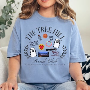 May include: Light blue t-shirt with a graphic print of the words "The Tree Hill Social Club" and various images from the TV show "One Tree Hill". The text "I don't want to be anything other than me" is printed below the graphic.