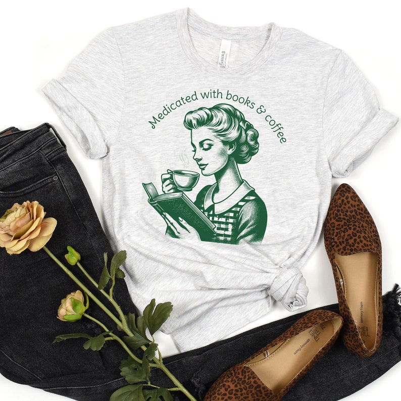 Op de afbeelding: Een grijs T-shirt met een groene illustratie van een vrouw die een boek leest en koffie drinkt. De tekst "Medicated with books & coffee" staat op het T-shirt gedrukt.