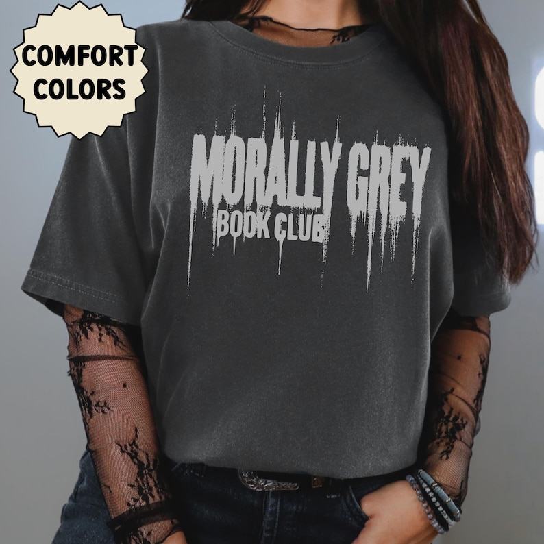 May include: Dark grey t-shirt with the text "MORALLY GREY BOOK CLUB" in distressed white lettering. The shirt has a crew neck and short sleeves. A small, cream-colored tag in the upper left corner reads "COMFORT COLORS".