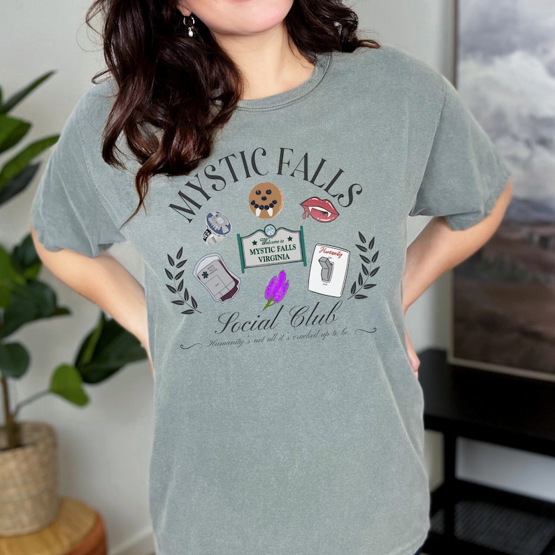 May include: A green t-shirt featuring a graphic print of the words "Mystic Falls Social Club" and various images, including a vampire fang, a blood bag, a cookie, and a sign that says "Welcome to Mystic Falls Virginia".