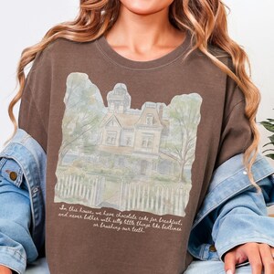 May include: A brown crewneck sweatshirt featuring a watercolor-style illustration of a large house with a picket fence. Below the house, the text reads: In this house, we have chocolate cake for breakfast, and never bother with silly little things like bedtimes or brushing our teeth.