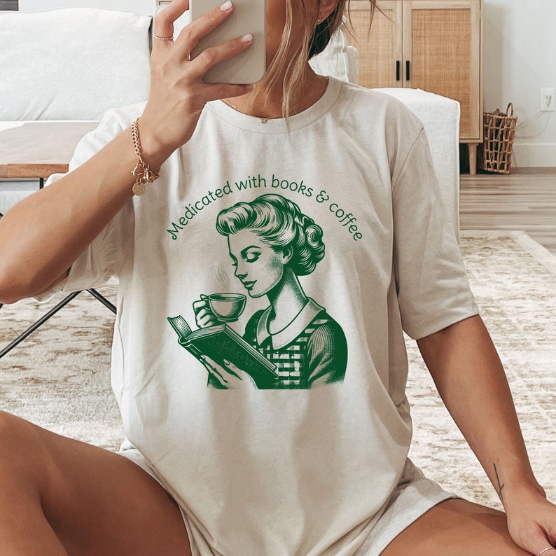 Op de afbeelding: Een beige T-shirt met een groene illustratie van een vrouw die een boek leest en koffie drinkt. De tekst "Medicated with books & coffee" staat op het T-shirt gedrukt.