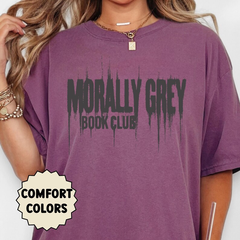May include: A purple t-shirt with the text "MORALLY GREY BOOK CLUB" in dark gray, dripping font. The shirt also features a "COMFORT COLORS" logo. The shirt is a solid color and has a crew neck.