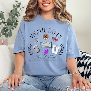 May include: A light blue t-shirt with a graphic design featuring the text "Mystic Falls Social Club" and various icons, including a vampire fang, a blood bag, and a cookie. The text "Welcome to Mystic Falls Virginia" is also included in the design.