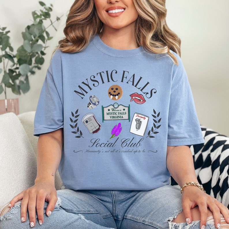 May include: A light blue t-shirt with a graphic design featuring the text "Mystic Falls Social Club" and various icons, including a vampire fang, a blood bag, and a cookie. The text "Welcome to Mystic Falls Virginia" is also included in the design.
