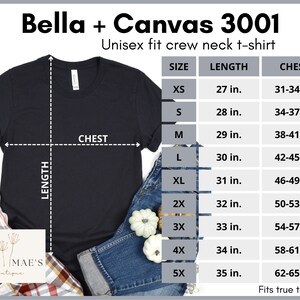 May include: A black unisex crew neck t-shirt with size chart showing measurements in inches. The chart includes sizes XS, S, M, L, XL, 2X, 3X, 4X, and 5X. The t-shirt is laid flat on a plaid surface with a pair of jeans and pumpkins.
