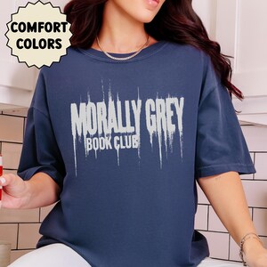May include: Navy blue t-shirt with the text "MORALLY GREY BOOK CLUB" in a distressed white font. The shirt also has a "COMFORT COLORS" logo in a cream-colored circle.