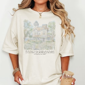 May include: A cream-colored short-sleeve t-shirt featuring a watercolor-style illustration of a Victorian house behind a white picket fence. Below the house, black cursive text reads: In this house, we have chocolate cake for breakfast.