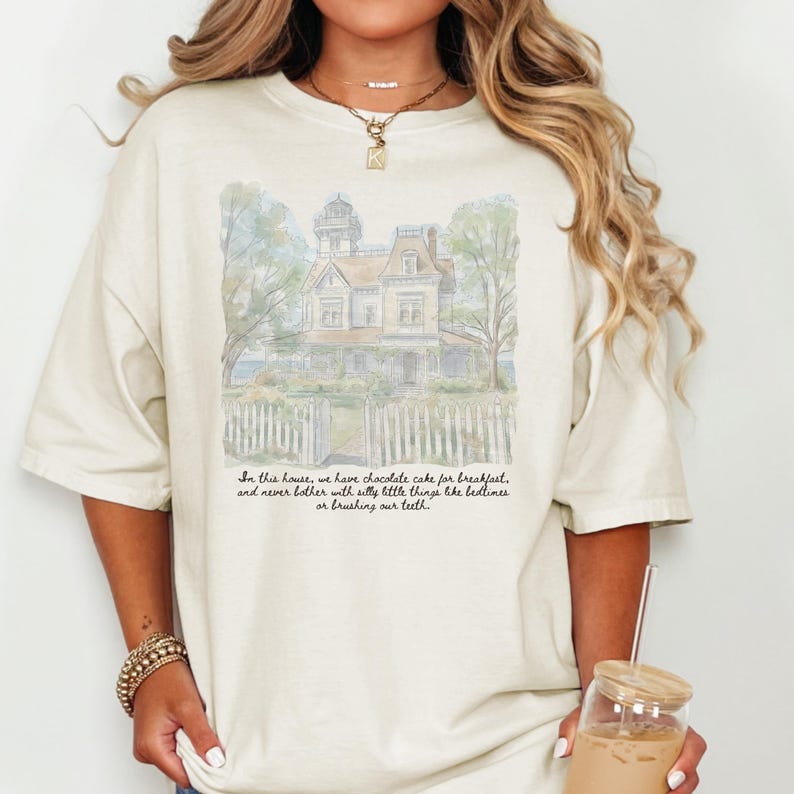 May include: A cream-colored short-sleeve t-shirt featuring a watercolor-style illustration of a Victorian house behind a white picket fence. Below the house, black cursive text reads: In this house, we have chocolate cake for breakfast.
